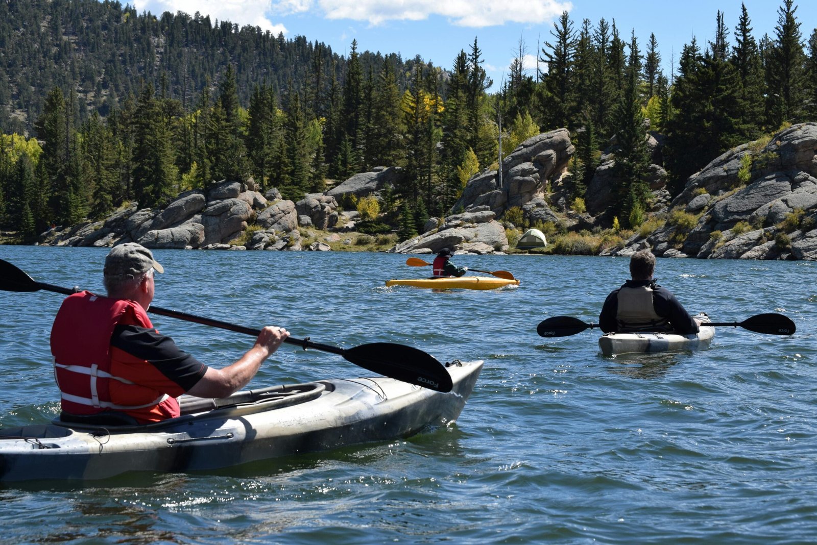 Three people kayaking on a picturesque lake surrounded by rocky terrain and evergreen trees.