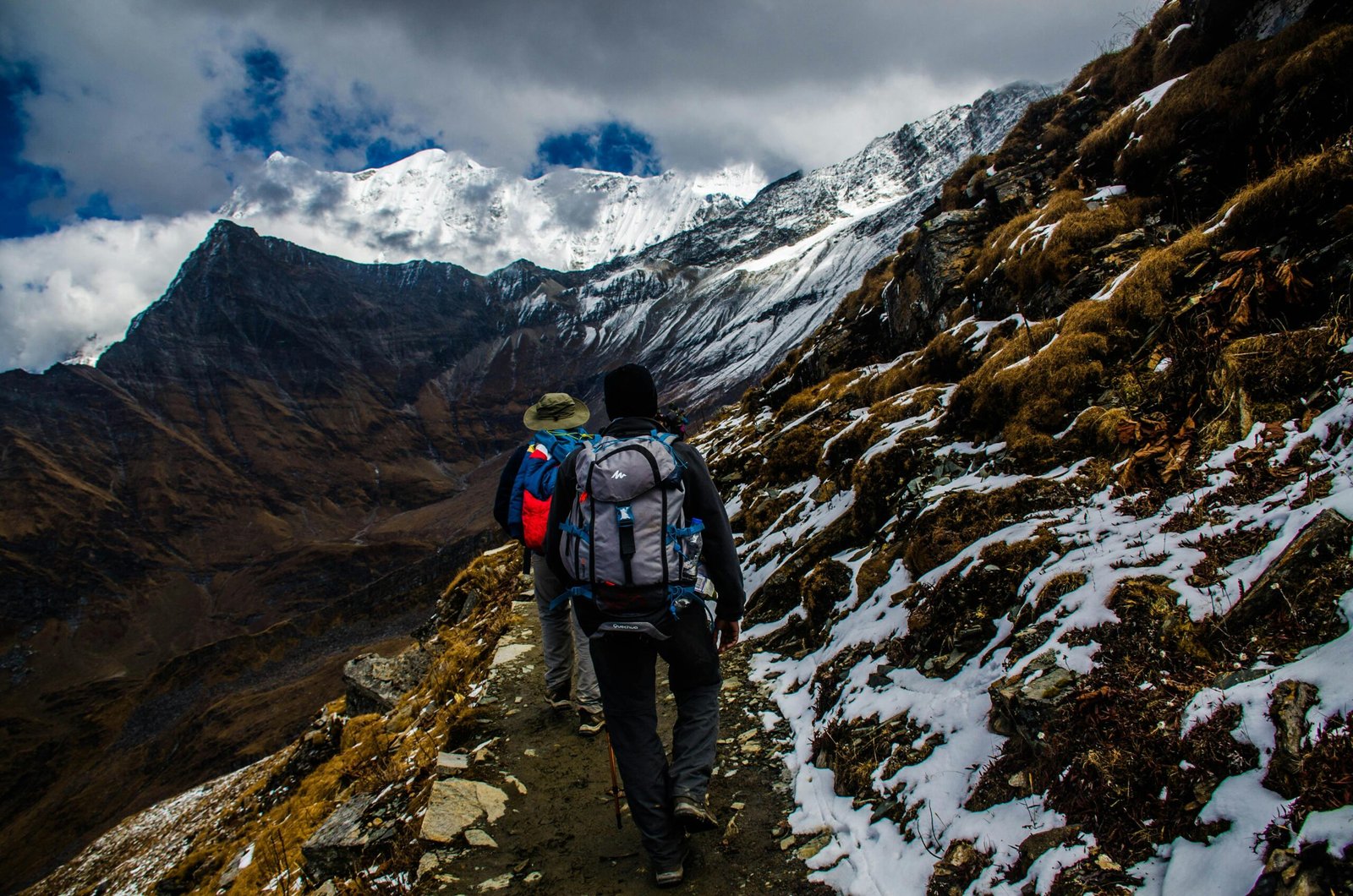 Two hikers navigating a snowy trail in rugged mountainous terrain under dramatic skies.